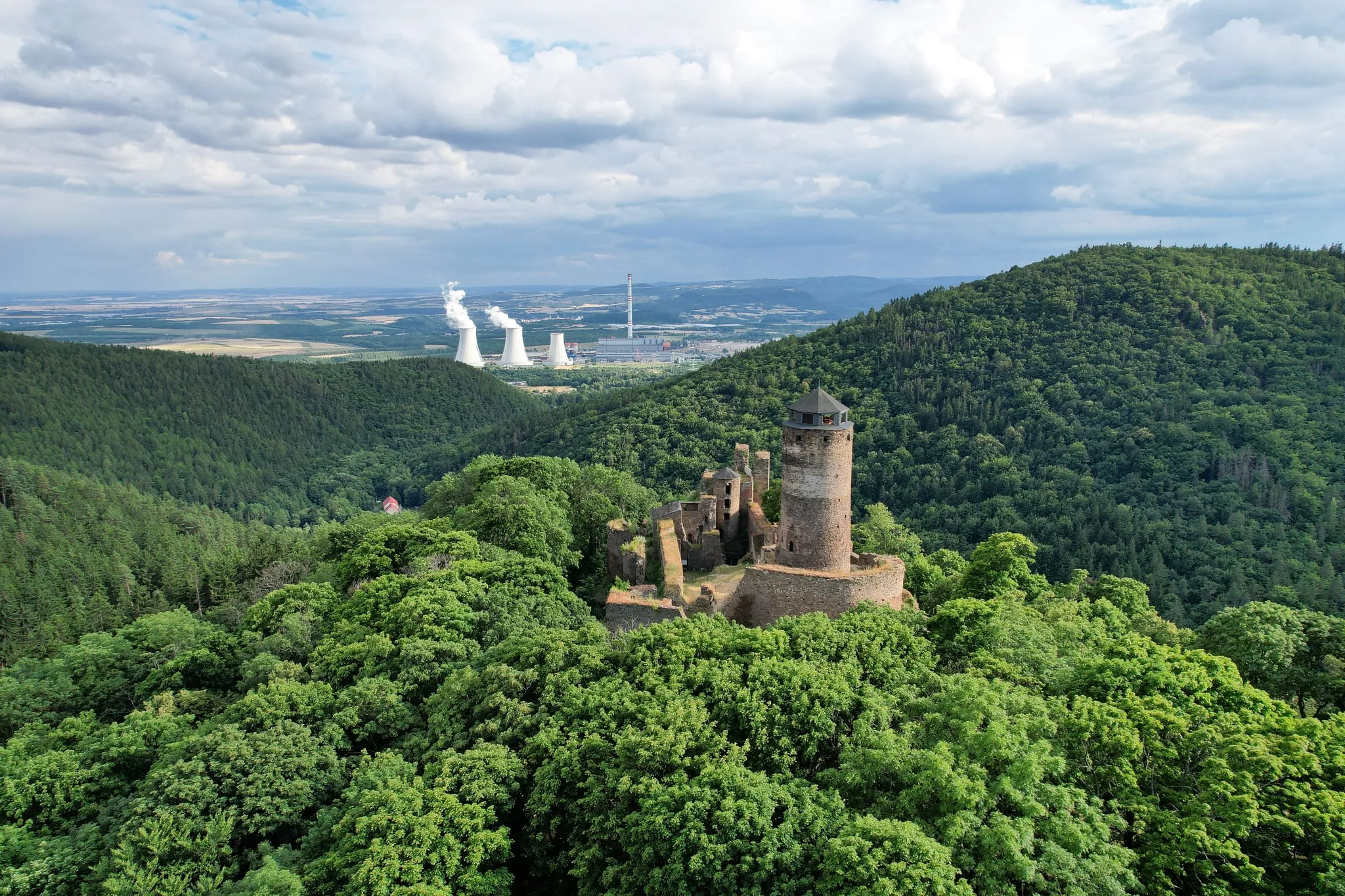 The ruins of Hasištejn Castle above dense deciduous forest, the cooling towers of a power station on the horizon