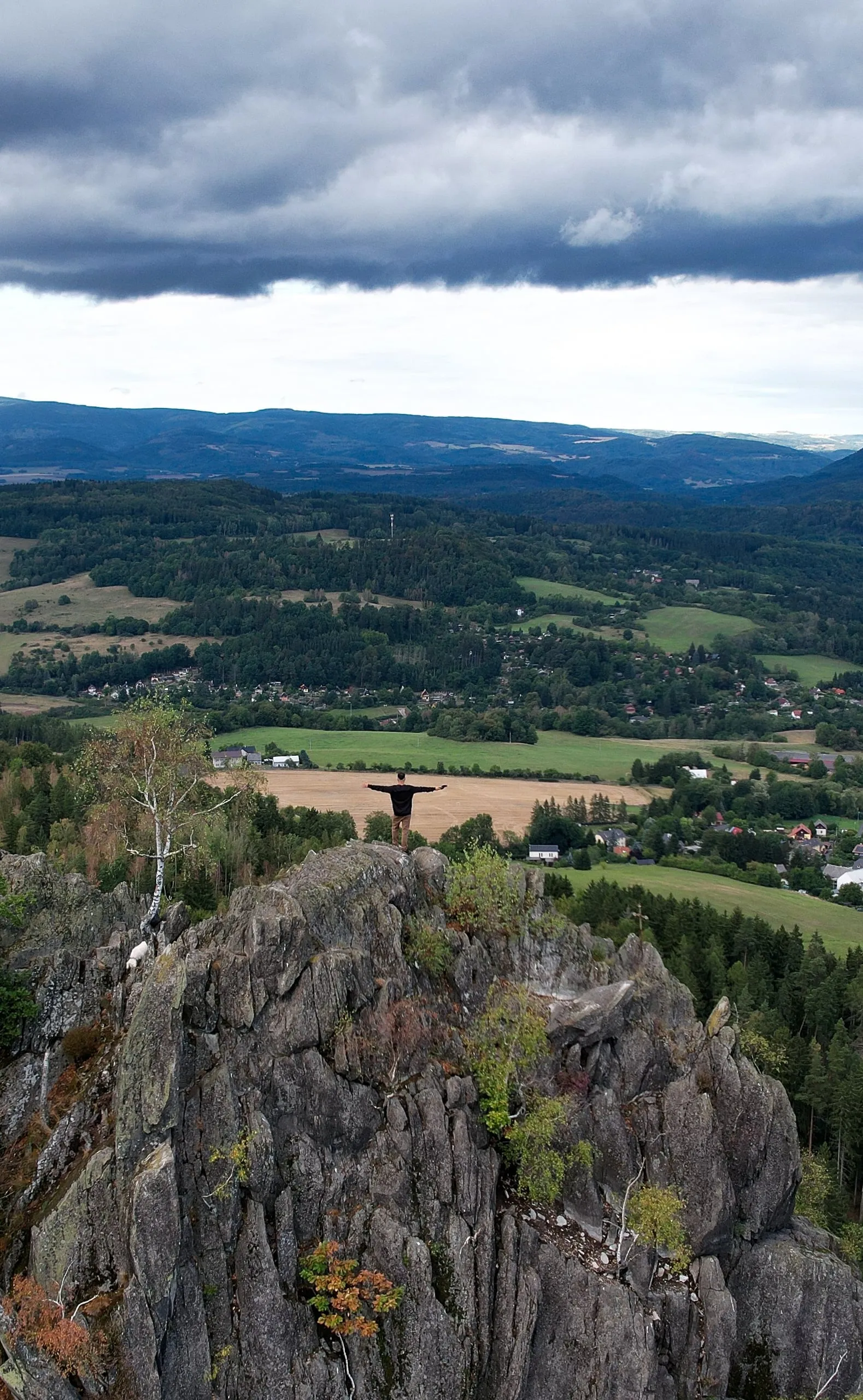A person with arms outstretched on top of the Šemnická skála rock, a valley with a village and wooded hills below under heavy clouds