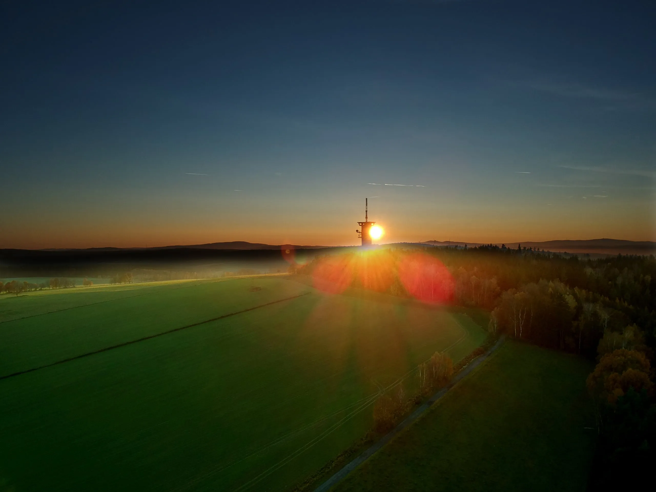 The sun setting right behind the lookout tower on Zelená hora, a green field in the foreground