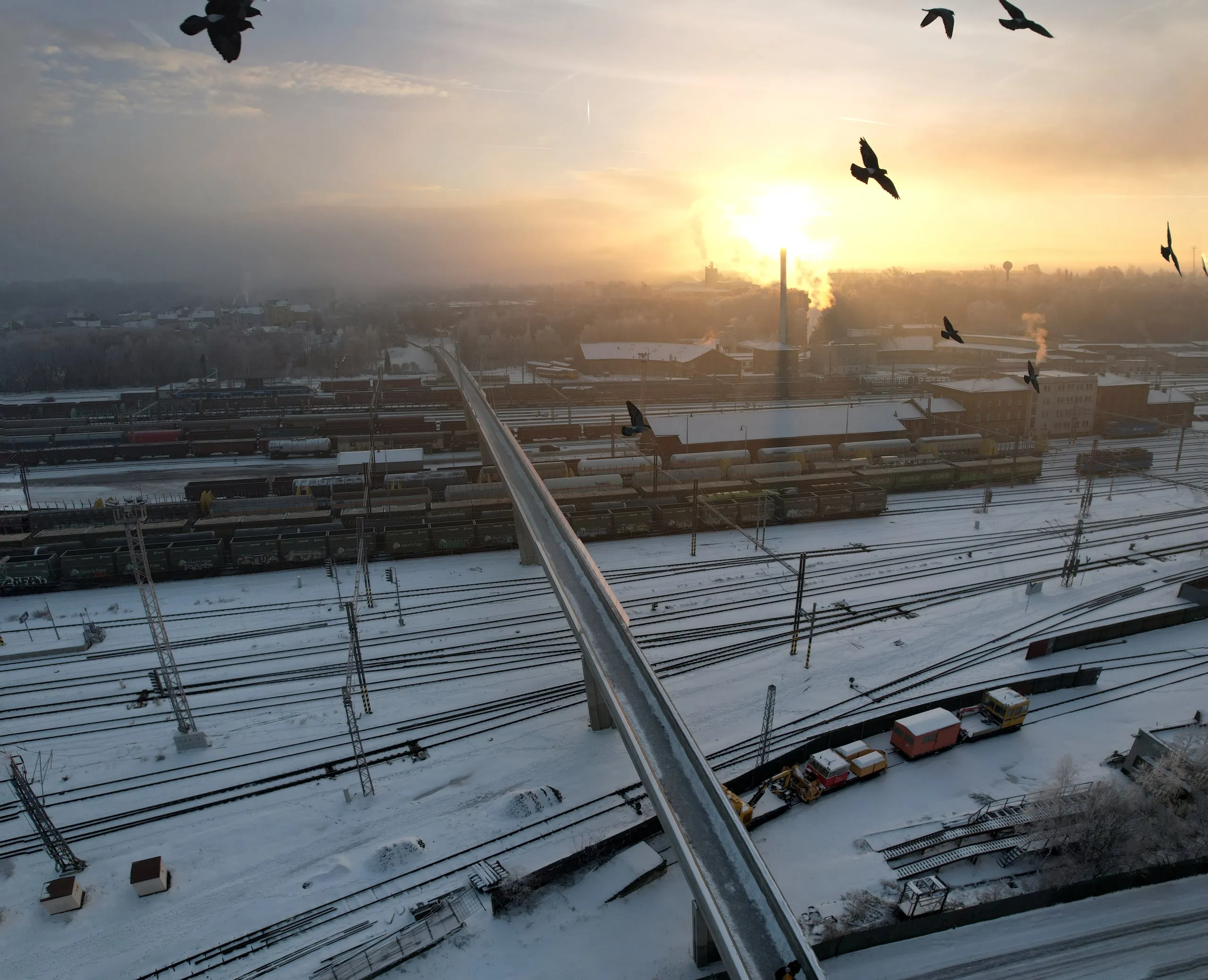 A snowy rail yard with a footbridge, birds flying against the sun